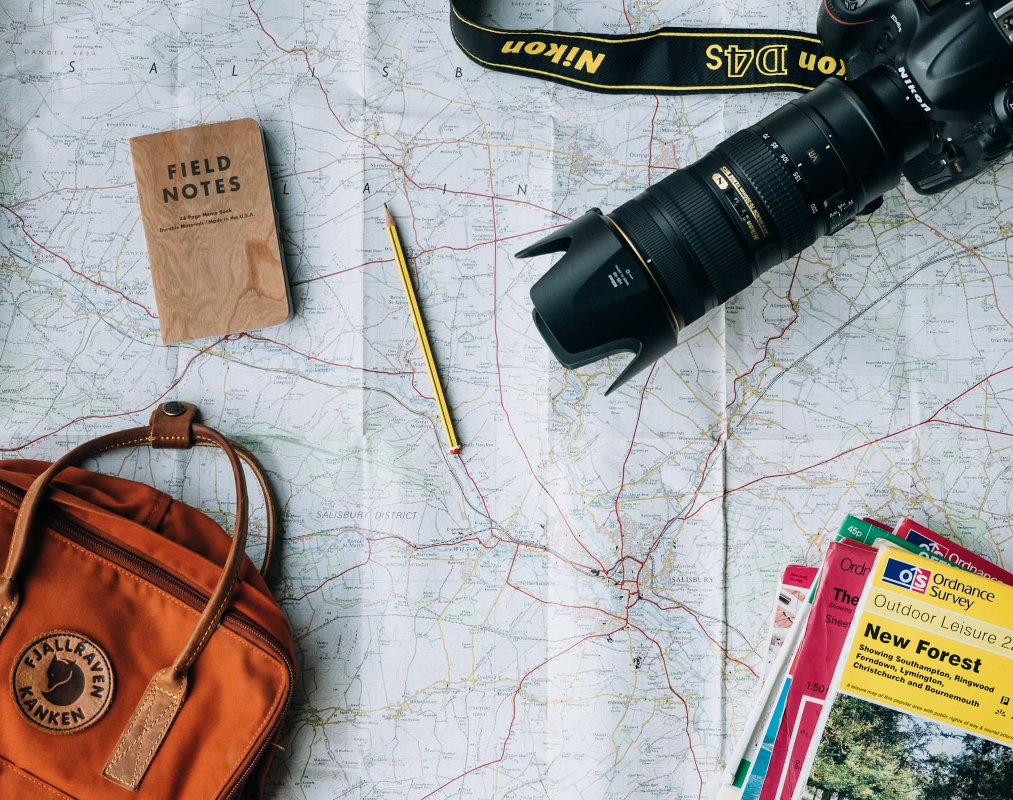 A paper map with field notes, a camera and a backpack laid out for trip planning